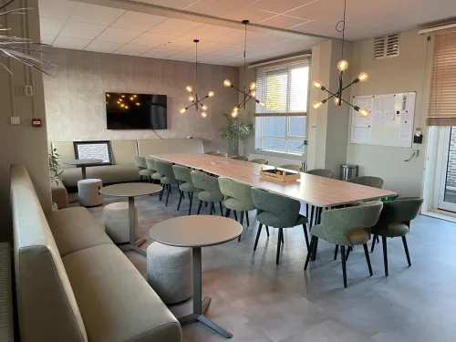 Lichte ontmoetingsruimte met lange vergadertafel, zithoek, ronde tafels en hanglampen in warme sfeer.