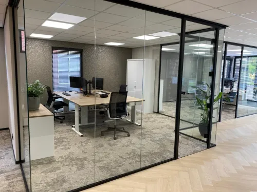 Strakke kantoorinrichting met glazen scheidingswanden, bureaus, stoelen en rustige kleurtonen.