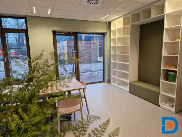 Lichte groepsruimte in kinderopvang met grote maatwerk wandkast, speelse zitnis en zicht op buiten via hoge ramen.