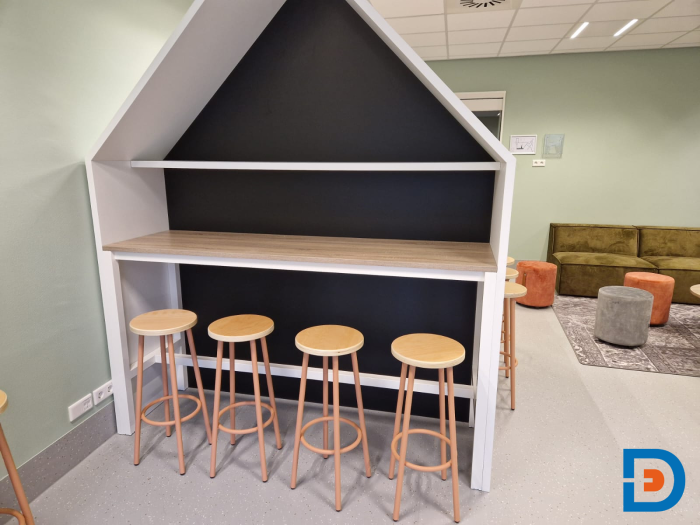 Speelhoek in de vorm van een huisje met hoge barkrukken, witte ombouw en krijtbordwand in de kinderopvang in Geesteren.