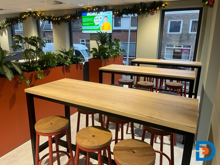 Moderne kantine met houten tafels, barkrukken en plantenbakken, ingericht voor informele ontmoetingen en groepsmaaltijden