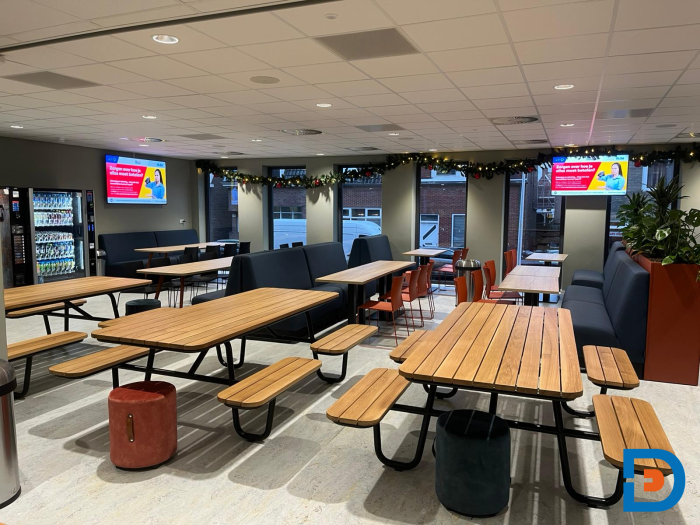 ROC Almelo kantine met een combinatie van treinbanken en picknickbanken, plantenbakken, kleurrijke poefjes en tafels met stoelen, ontworpen voor informele eet- en ontmoetingsmomenten.